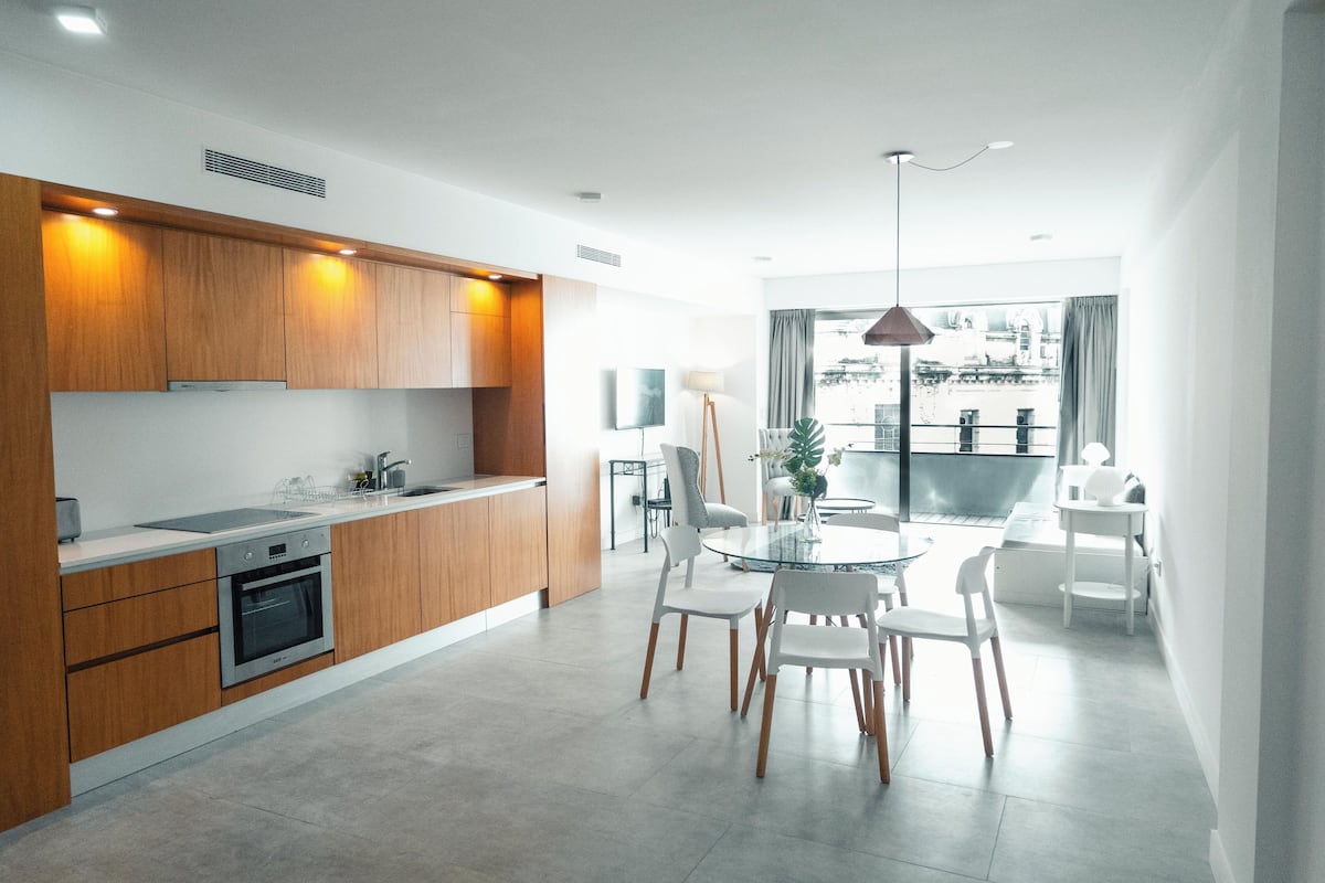A modern kitchen with wooden cabinets, a built-in oven, and a dining area with a glass table and white chairs.
