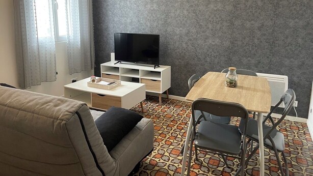 A living room with a sofa, a TV on a white stand, a small table with a vase, and a patterned floor. - 1 Bedroom Apartment in BÃ©ziers (Béziers)