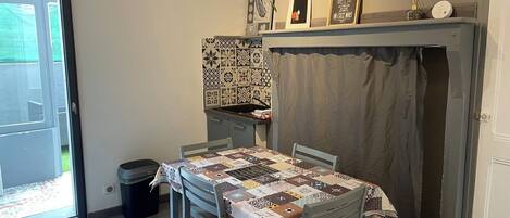 A dining area with a table covered by a patchwork tablecloth, a patterned wall hanging above the table, and a small shelf with decorative items.