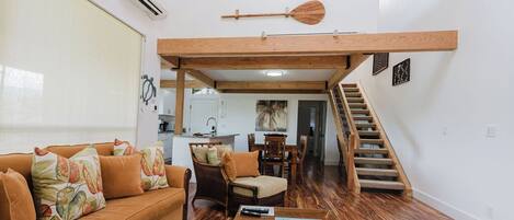 A spacious living area with a high ceiling, wooden beams, a sofa with patterned cushions, a dining table, and a staircase leading to an upper level.