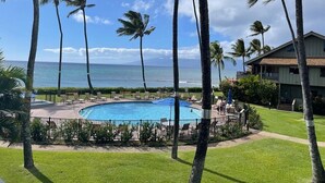 A pool area with palm trees, a view of the ocean, and a building in the background. - Ocean View, Beach Front 2bd 2ba unit less than a minute walk to the beach! (Lahaina)