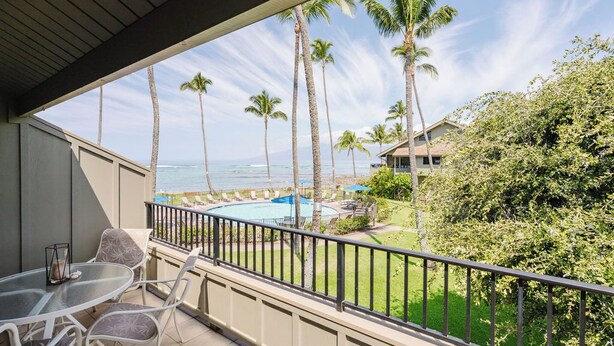 A balcony with a glass table and chairs, overlooking a pool and palm trees, with a view of the ocean and a building in the background. - Ocean View, Beach Front 2bd 2ba unit less than a minute walk to the beach! (Lahaina)
