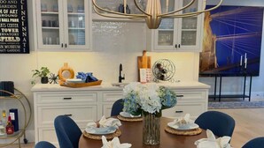 Un coin repas avec une table ronde dressée pour six personnes, entourée de chaises recouvertes d’un tissu bleu. Un lustre est suspendu au-dessus, et un vase rempli de fleurs blanches se trouve sur la table.