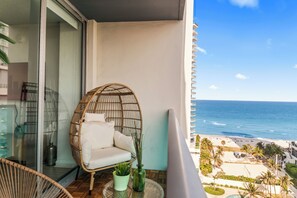 Un balcon avec un fauteuil en osier, une petite table avec une plante, et une vue sur la plage et l’océan.