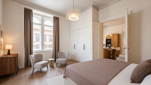 A hotel room with a large bed, two armchairs, a small table, a desk with a lamp, a chandelier, and a view of the city through the window.