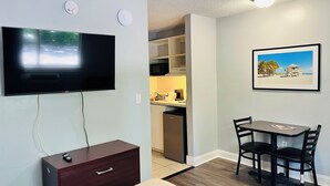 Ein kompakter Wohnbereich mit Flachbildfernseher an der Wand, einem kleinen Esstisch mit Stühlen, einer dunklen Holzschrankwand und einem durch eine offene Tür einsehbarer Küchenbereich.