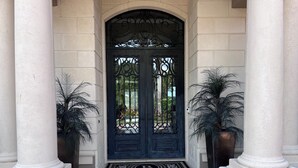 Una puerta doble negra con ornamentación metálica detallada, flanqueada por columnas blancas y plantas en macetas.