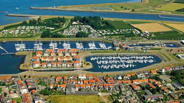 Vista aérea de un puerto deportivo costero con numerosas embarcaciones amarradas, rodeado de edificios residenciales y campos verdes.