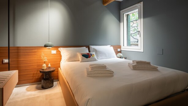 Une chambre moderne avec un grand lit, une tête de lit en bois, une table de chevet avec une lampe et une fenêtre donnant sur un paysage verdoyant.