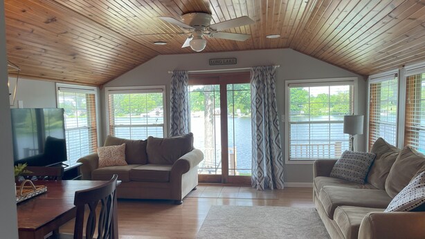 A spacious living room with a wooden ceiling, a large window overlooking a lake, a television, and a cozy seating area with a sofa and a chair. - Cozy Lakefront Cottage (Colon)
