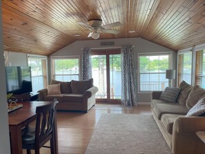 Een ruime woonkamer met een houten plafond, een groot raam met uitzicht op een meer, een televisie en een gezellige zithoek met een bank en een fauteuil.