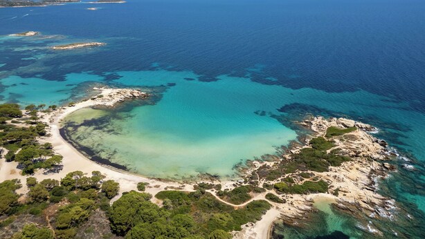 An aerial view of a coastal area with clear turquoise waters, sandy beaches, and rocky terrain with sparse vegetation.