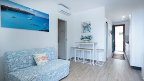 Ett modernt vardagsrum med en mönstrad soffa, ett litet vitt bord med två pallar, en tavla som föreställer ett kustlandskap och utsikt in i köket.