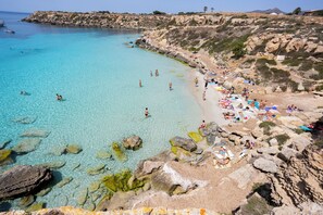 Ein Küstenstrand mit klarem türkisblauem Wasser, felsigem Ufer und badenden Personen.