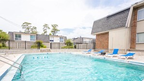 Una piscina rectangular con agua azul cristalina, rodeada por un deck de baldosas. Hay sillas reclinables azules a lo largo de la piscina.
