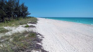 En strand med vit sand, kristallklart blått vatten och sparsam växtlighet.