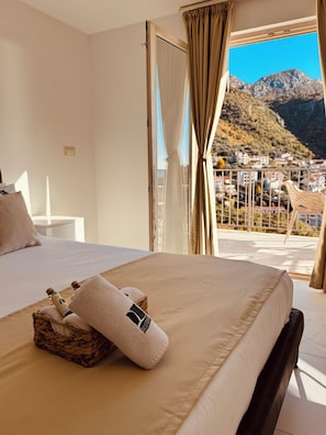 Quarto de hotel com uma cama, uma mesa pequena e vista para uma paisagem montanhosa através de uma varanda aberta.