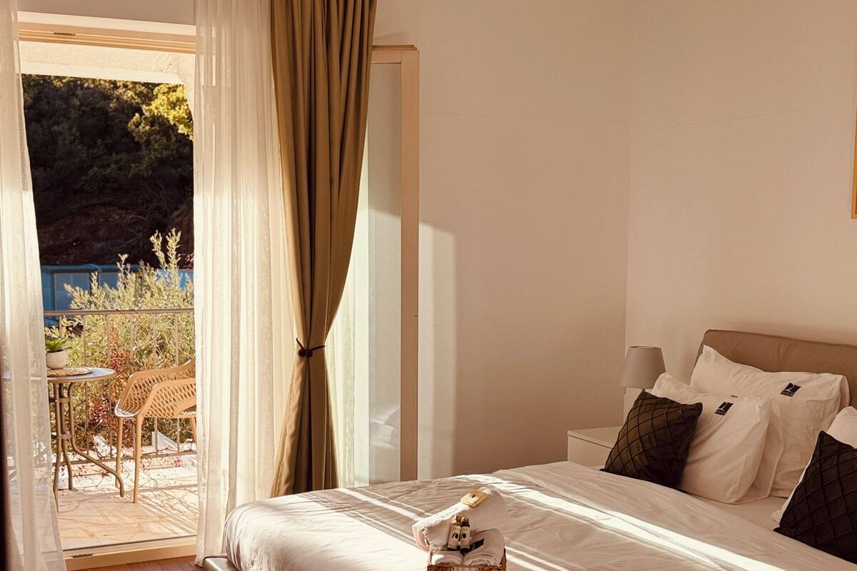 A bedroom with a bed, a bedside table, a lamp, and a view of an outdoor area with a chair and plants.