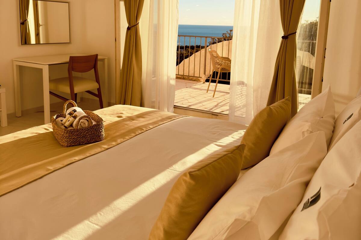 A hotel room with a bed, a desk, and a chair. There is a view of the sea through the curtains.