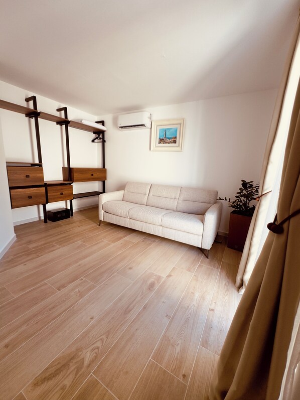 Quarto com piso de madeira, um sofá bege, uma escrivaninha de madeira com gavetas, um cabideiro suspenso, um aparelho de ar condicionado e um quadro na parede.