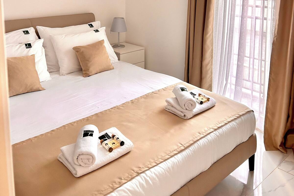 A hotel room with a neatly made bed, a bedside table with a lamp, and a window with curtains.