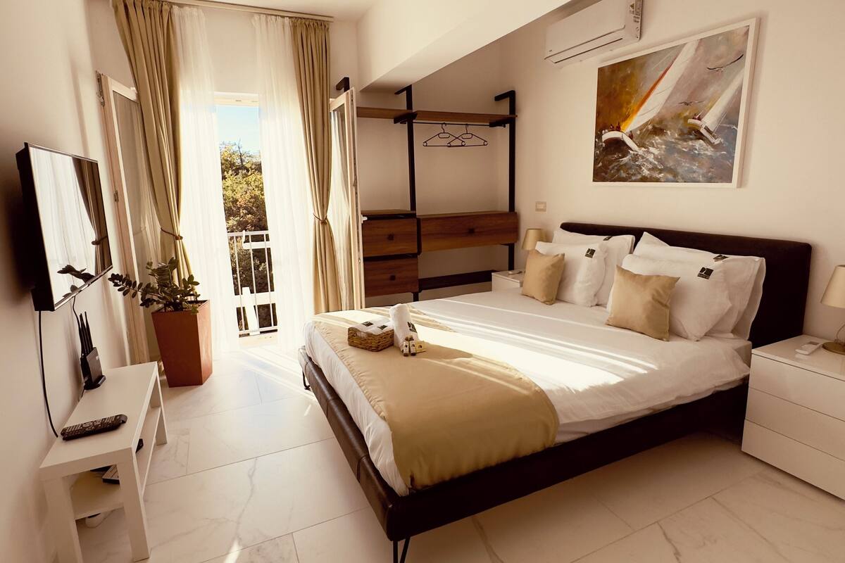 A modern hotel room with a large bed, a bedside table, a TV, and a balcony with curtains.