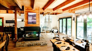 Una sala de estar acogedora con chimenea, vigas de madera, revestimiento de piedra para la chimenea, un comedor con una mesa dispuesta para cuatro personas y grandes ventanales que ofrecen vistas al exterior.