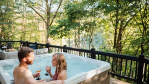 Un jacuzzi con dos personas sosteniendo bebidas, rodeado por una terraza de madera y árboles.