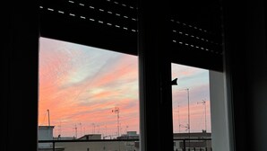 Une vue à travers une fenêtre ornée d’une grille métallique, donnant sur un toit avec des bâtiments et des antennes.