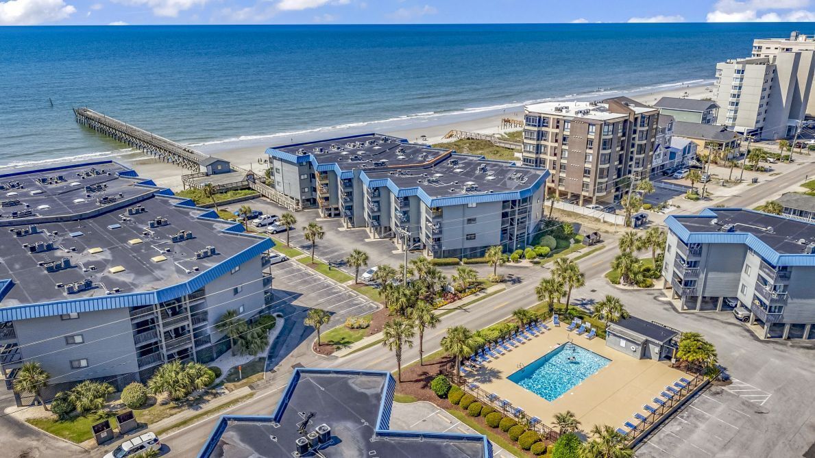 A coastal hotel with multiple buildings, a parking lot, a swimming pool, and a pier in the background.