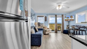 Eine moderne Küche mit einem Edelstahl-Kühlschrank, ein Essbereich mit Tisch und Stühlen, ein blaues Sofa und ein Blick auf den Ozean durch große Fenster.