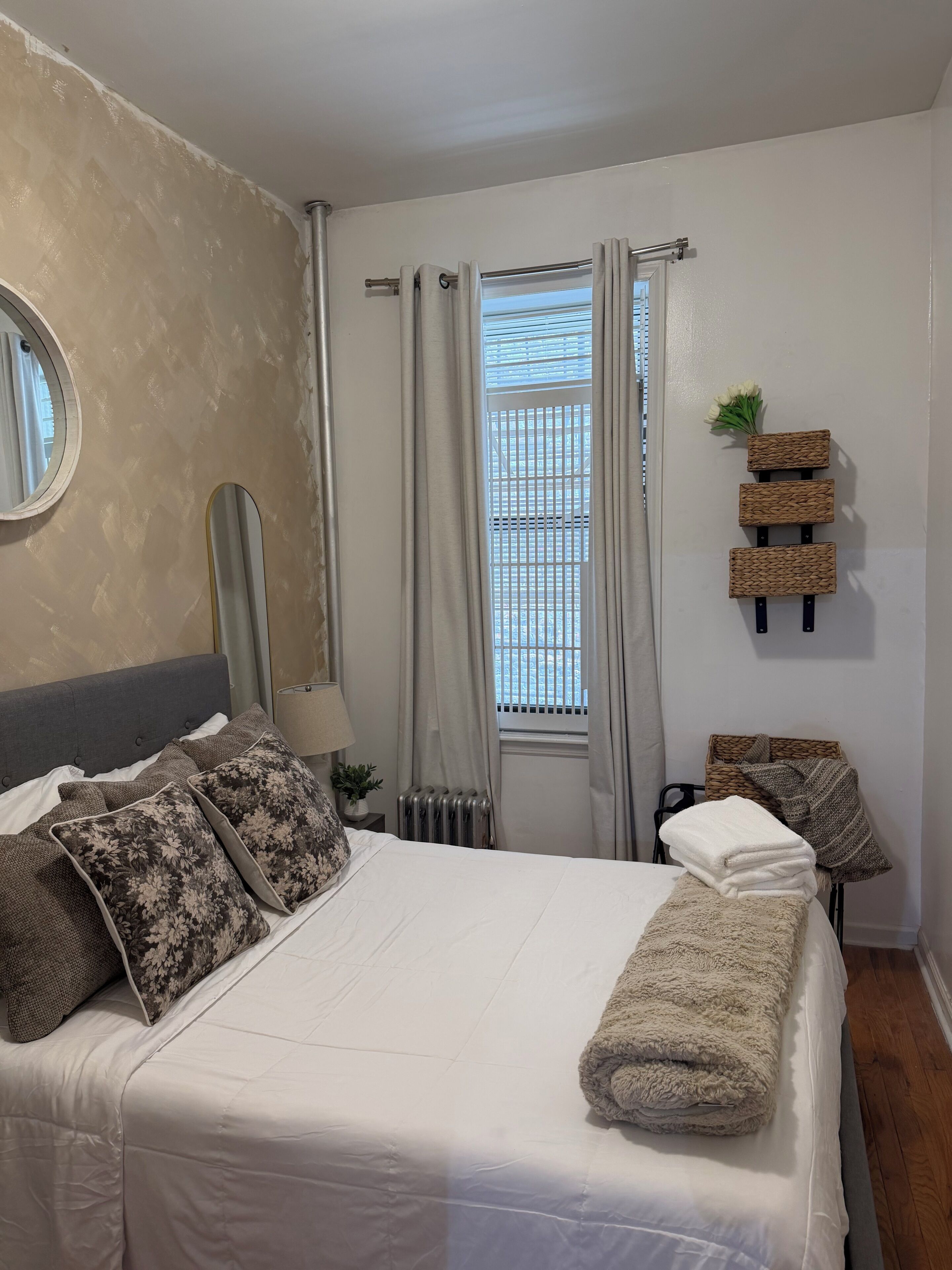 A neatly made bed with a white comforter and a rolled towel, a round mirror on the wall, a radiator, and a window with curtains.