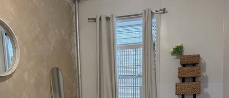 A neatly made bed with a white comforter and a rolled towel, a round mirror on the wall, a radiator, and a window with curtains.
