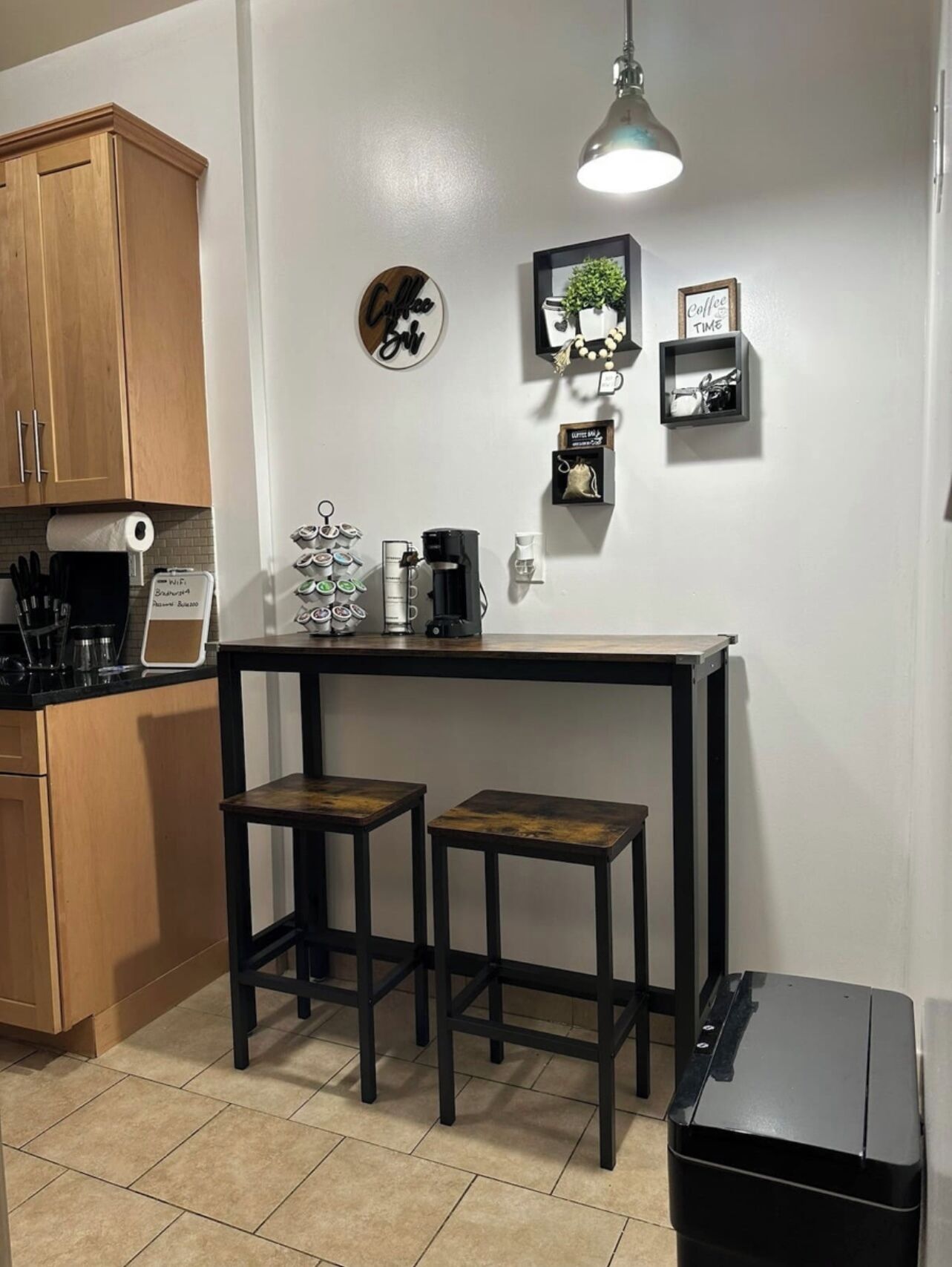 Una cocina pequeña con armarios de madera, una zona de café y un cubo de basura negro.
