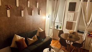 Una sala de estar con un sofá verde oscuro, varios cojines, una mesa de centro redonda y una zona de comedor con una mesa pequeña y sillas.