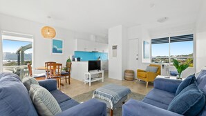 A modern living room with a sofa set, a dining area with a table and chairs, a kitchen with white cabinets, and large windows offering a view of the outdoors. - 4-53 Forsters Bay Road, Narooma - Blue Water Villas (Narooma)