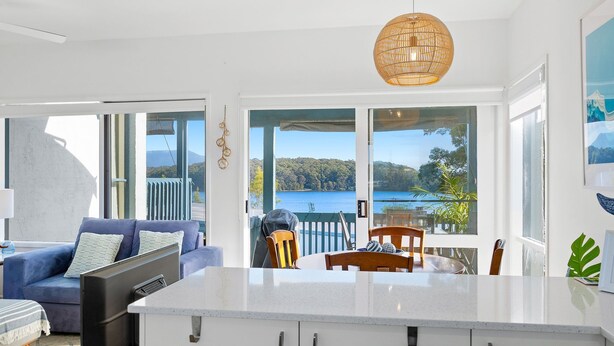 A modern kitchen with a large island, a dining area with a view of a lake, and a living room with a sofa and a TV. - 4-53 Forsters Bay Road, Narooma - Blue Water Villas (Narooma)