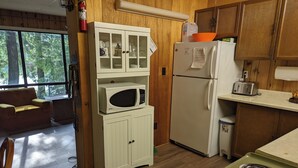 A compact kitchen with a microwave, refrigerator, and stove. There is a wooden cabinet with glass doors, a wooden floor, and a window providing a view of trees.