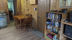 A wooden-paneled room with a dining table and chairs, a bookshelf filled with books and DVDs, and a kitchen area with a stove and oven.