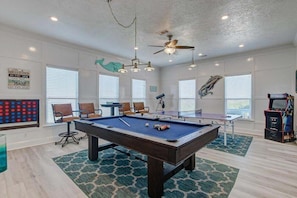 Une table de billard dans une pièce aux murs blancs, avec un ventilateur de plafond et un jeu de fléchettes.