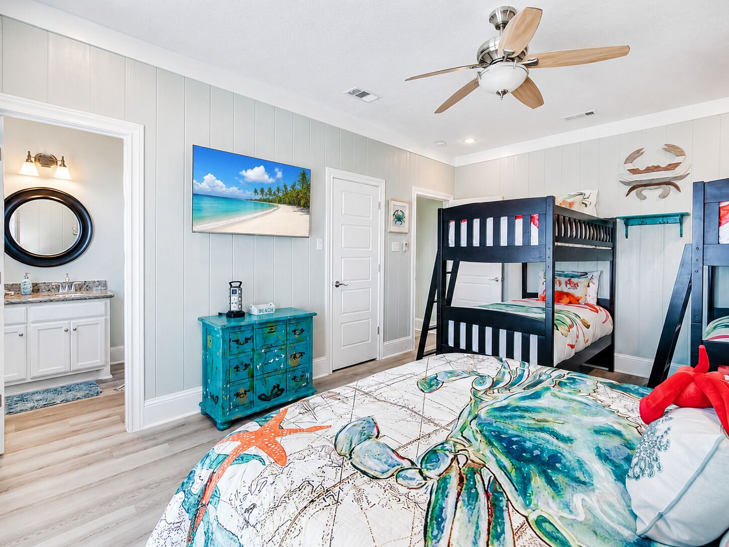 A bedroom with a bunk bed, a ceiling fan, a bathroom with a vanity, and a beach-themed wall art.