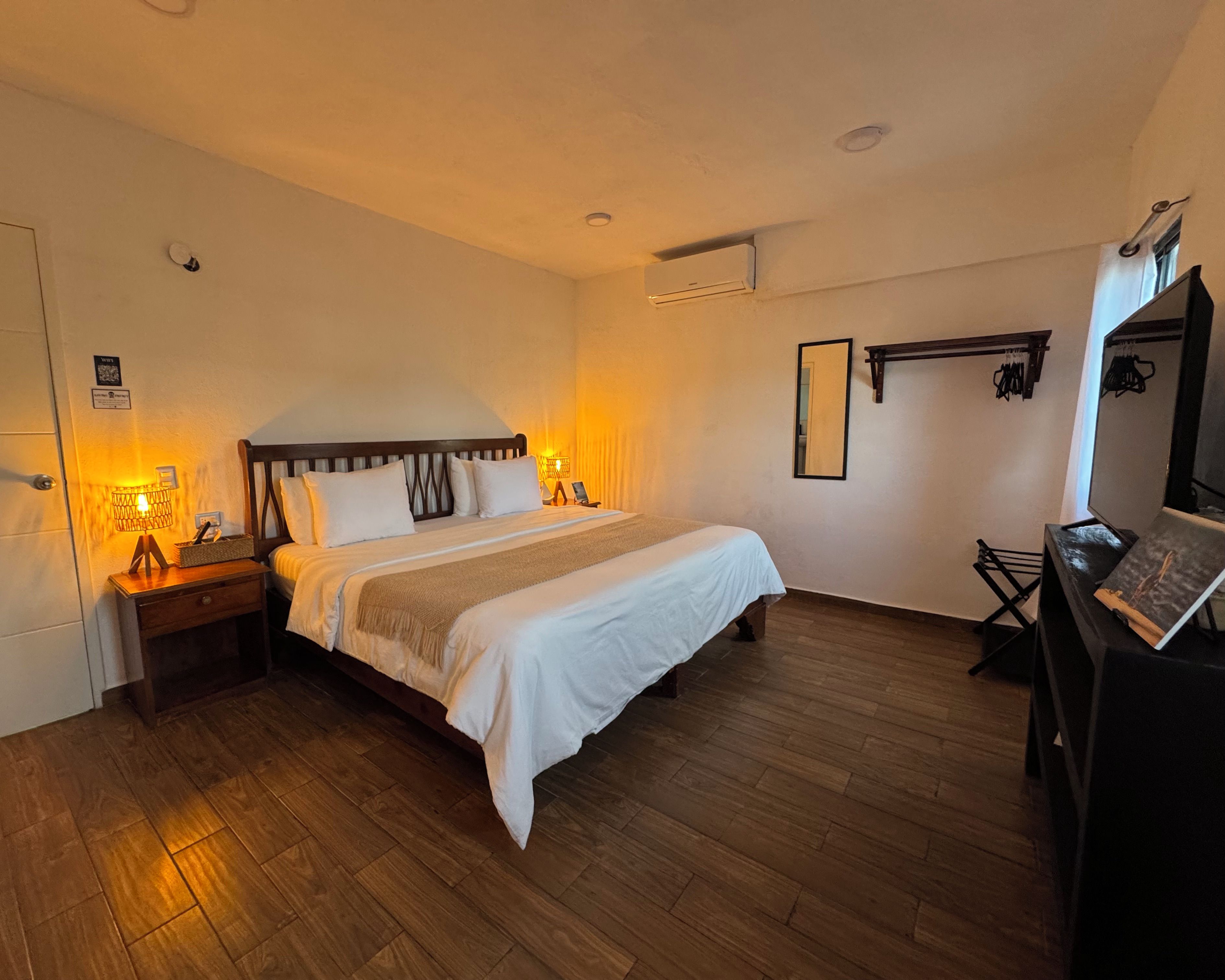A hotel room with a bed, bedside tables, an air conditioning unit, a mirror, and a desk with a chair.