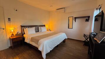 A hotel room with a bed, bedside tables, an air conditioning unit, a mirror, and a desk with a chair.