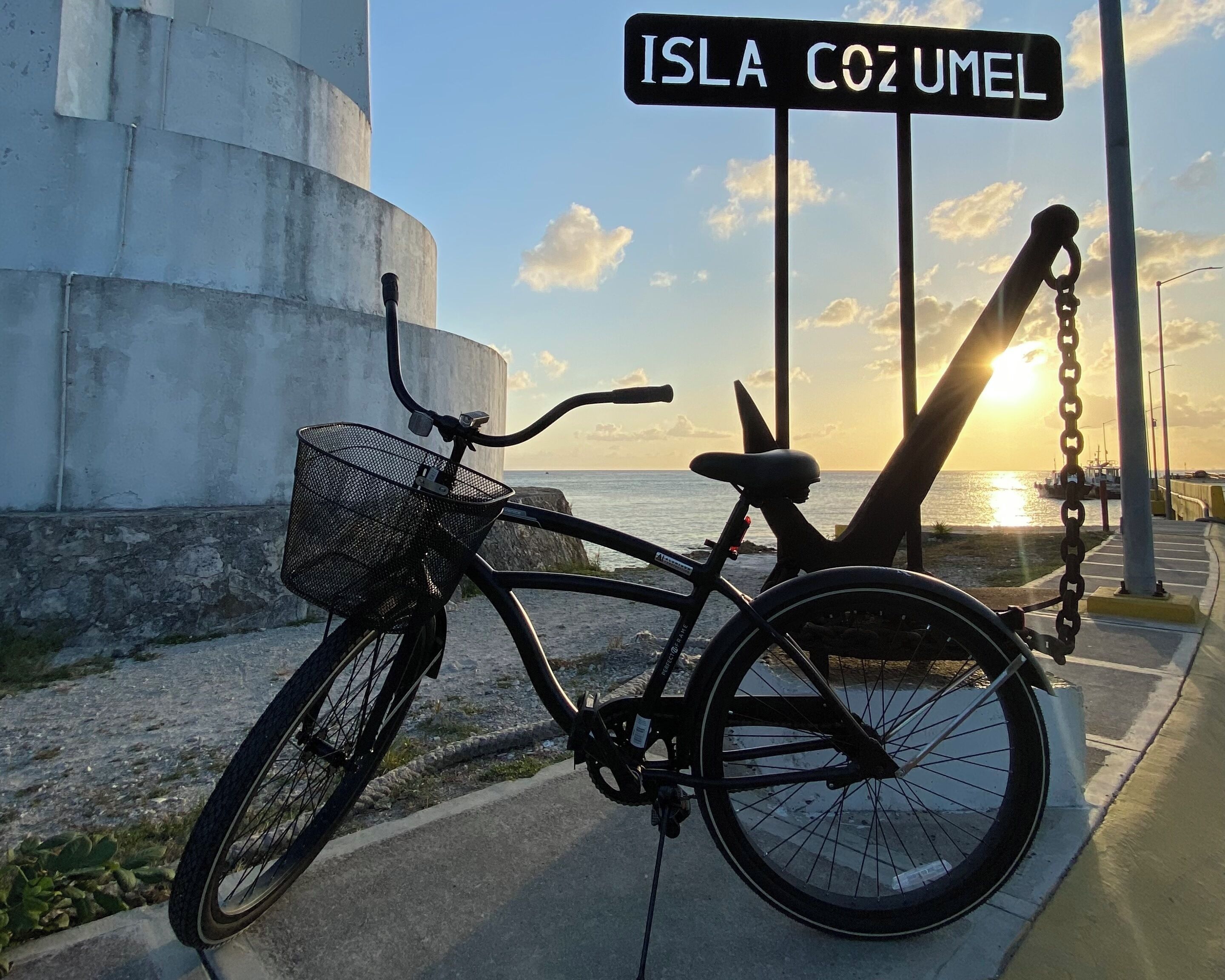 Una bicicleta estacionada cerca de un letrero que dice "ISLA COZUMEL", con un atardecer de fondo.