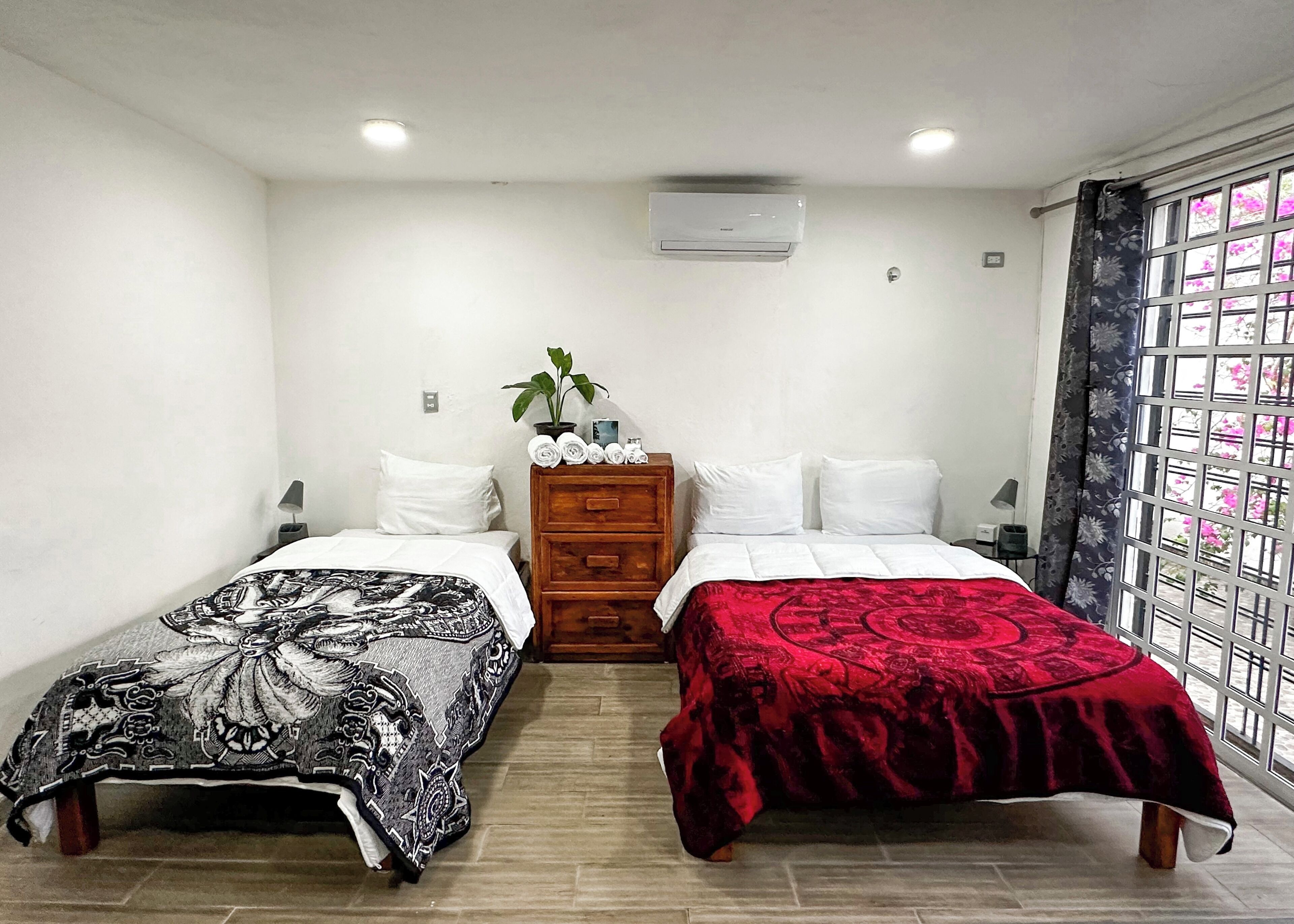 A hotel room with two beds, a wooden dresser, a potted plant, and a window with a view of pink flowers.