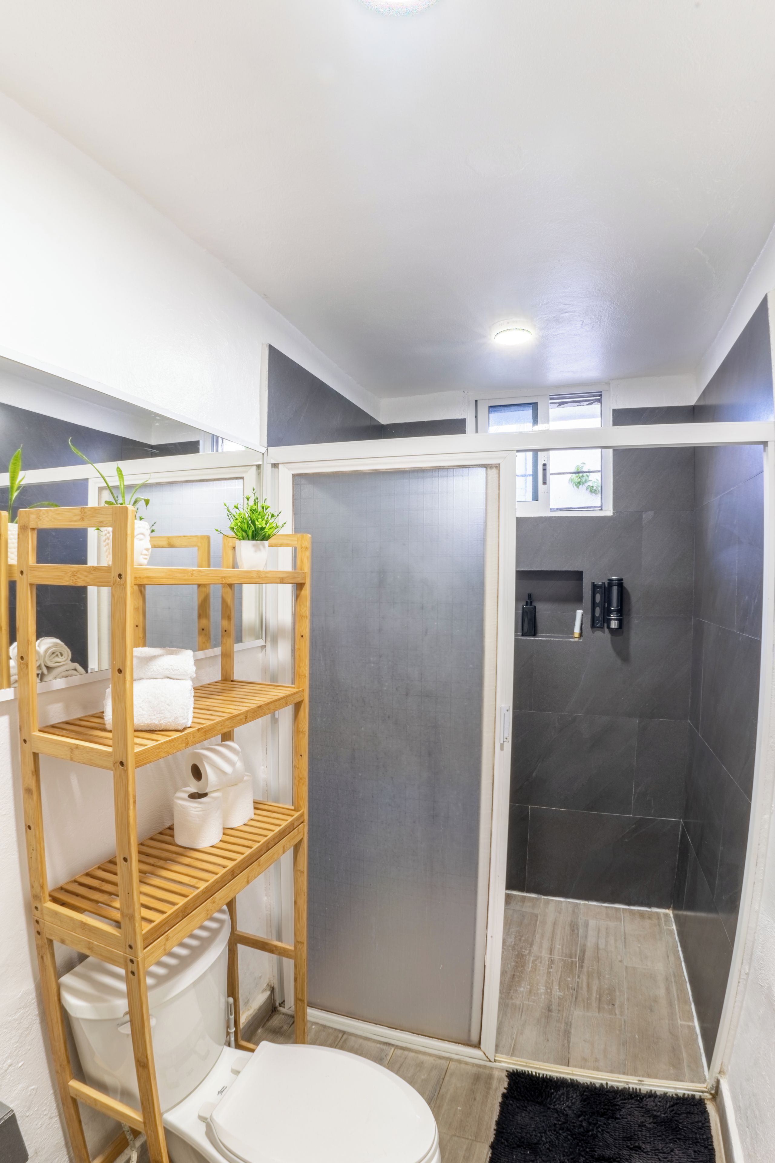 Et badeværelse med bruser, toilet, en træhylde med håndklæder og en lille plante.