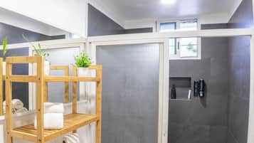 Et badeværelse med bruser, toilet, en træhylde med håndklæder og en lille plante.