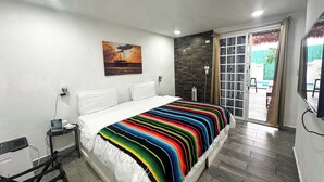 A hotel room with a bed, a small table with a lamp, a painting on the wall, and a view of the outside. - La Perle Noire (Cozumel)