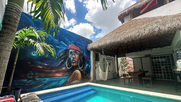 A swimming pool with a mural of a pirate and a ship, surrounded by palm trees and a thatched-roof structure. - La Perle Noire (Cozumel)