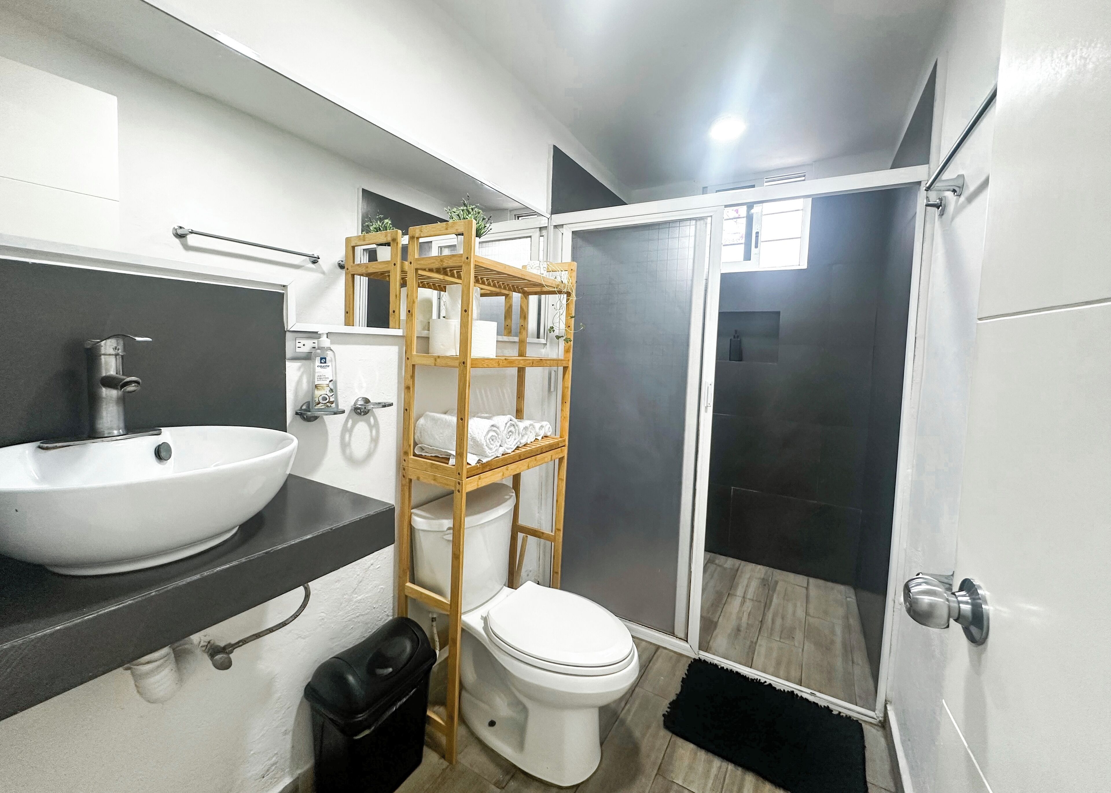 Une salle de bain moderne avec un lavabo, des toilettes et une douche, le tout de couleur blanche. On y trouve une étagère en bois où sont rangées des serviettes et des articles de toilette.